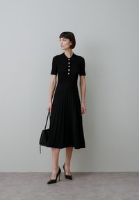 Must volditud kleit lühikeste varrukatega, krae juures nööpide detail, sobitatud musta käekoti ja kõrgete kontsadega kingadega.
