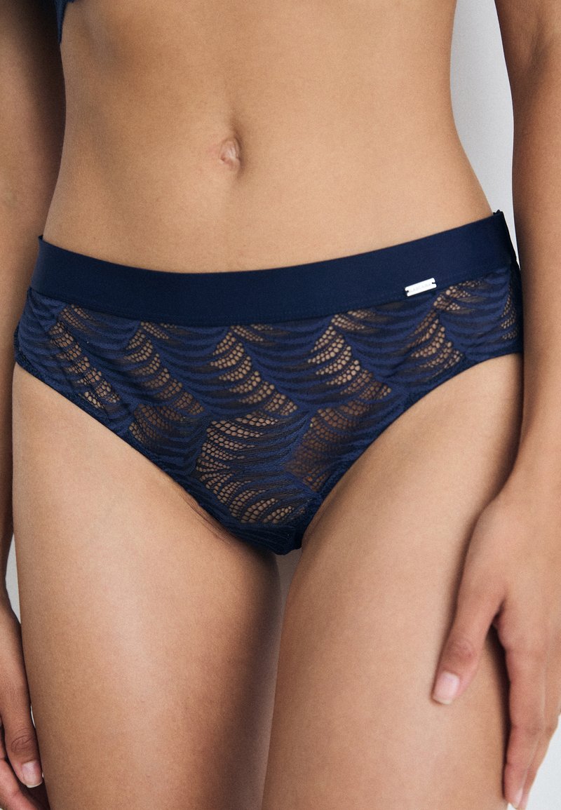 Parte de baixo de biquíni de renda azul-marinho com um padrão ondulado e uma cintura grossa e sólida, adornada com um pequeno detalhe metálico.