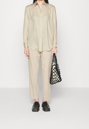 Béžová košile na zapínání s límečkem, v kombinaci s světle béžovými kalhotami a černými nazouvacími botami. Drží černou a krémovou tkanou tašku.