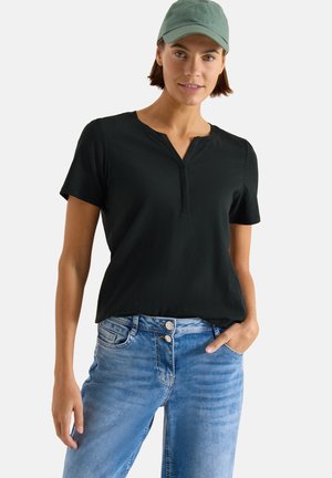 Femme portant une casquette verte, un t-shirt henley noir à manches courtes et un jean bleu clair à boutons doubles, debout avec une main dans la poche.