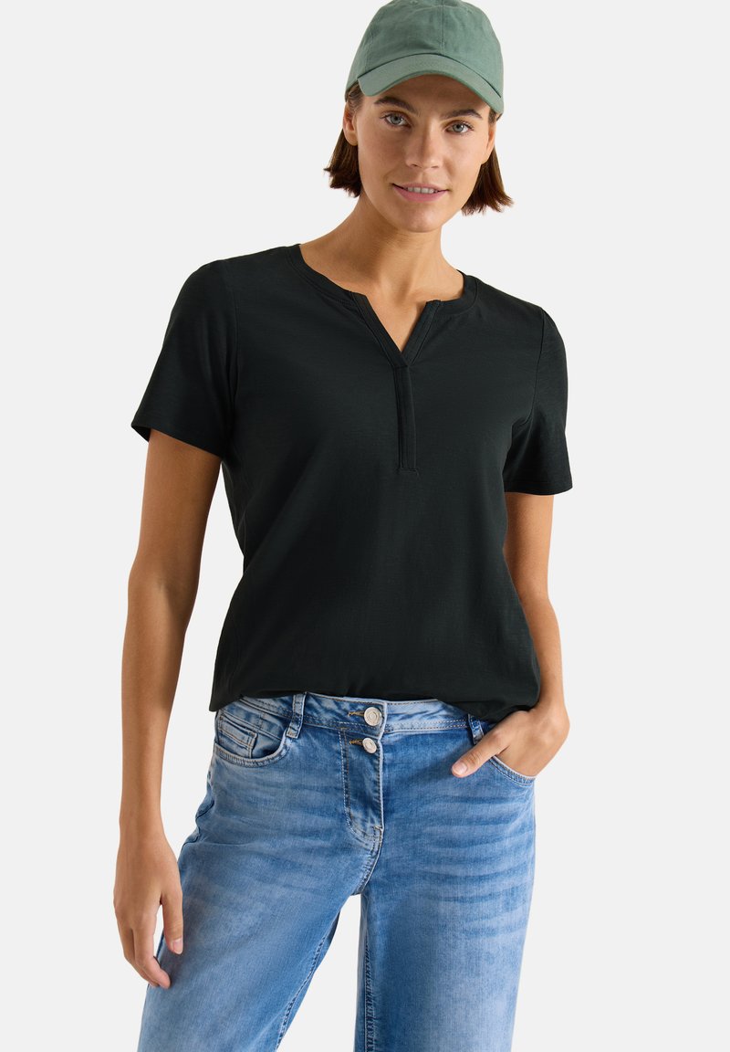 Femme portant une casquette verte, un t-shirt henley noir à manches courtes et un jean bleu clair à boutons doubles, debout avec une main dans la poche.