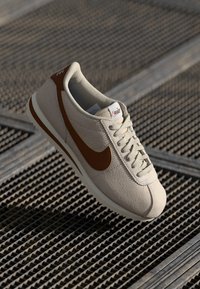 Bežna športna čevlja z teksturirano površino in rjavimi poudarki, ki se ponaša s klasičnim dizajnom, okroglo konico in tankimi vezalkami. Prikazano je Nikeovo blagovno ime.