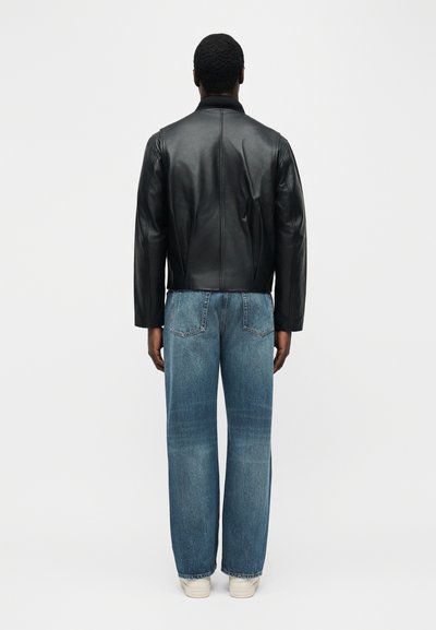 Άνδρας με την πλάτη, φοράει μαύρο δερμάτινο τζάκετ, μπλε τζιν κανονικής γραμμής και λευκά αθλητικά παπούτσια, μπροστά σε απλό λευκό φόντο.