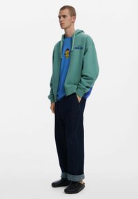 Zelena zip-hoodica preko plave majice s grafikom, u paru s širokim tamnim trapericama i crnim slip-on cipelama s prebačenim rubovima.