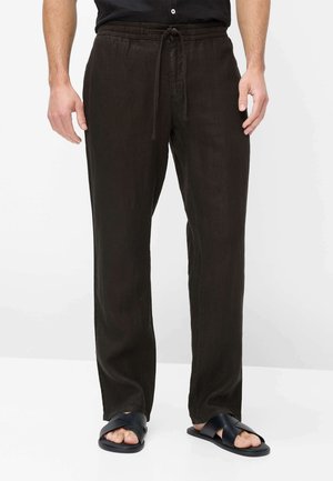 Mand iført løse sorte snorebukser og sorte slidesandaler, stående mod en ensfarvet hvid baggrund.