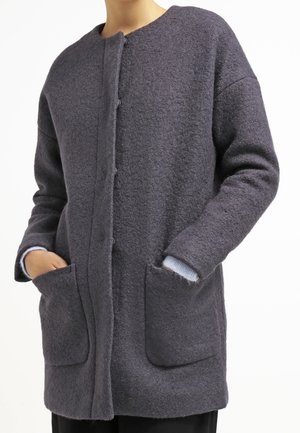 Femme portant un long manteau gris foncé texturé avec de grandes poches avant, les mains partiellement dans les poches, et une chemise bleu clair en dessous.