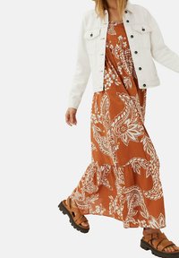 Maxikjole i rustfarve med hvide blomstrede paisley-mønstre, lagdelt flæsekant, kombineret med en hvid denimjakke og brune sandaler med remme.