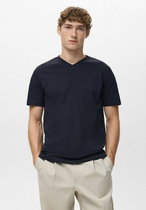 Jonge man die een donkerblauw V-hals T-shirt met korte mouwen en licht beige broek draagt, staand met handen in de zakken tegen een effen witte achtergrond.