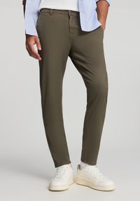Pantaloni verde oliva con vestibilità slim, realizzati in materiale liscio. Indossati con sneakers bianche, in piedi davanti a uno sfondo neutro.