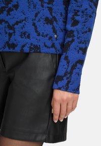 Niebieski sweter o teksturze z czarnym wzorem; posiada srebrny guzik jako akcent. Zestawiony z czarnymi skórzanymi szortami, które podkreślają gładkie wykończenie.