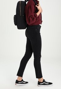 Zwarte rugzak met gouden ritsen, gedragen over een bordeauxrode cropped blouse, gecombineerd met zwarte distressed jeans en zwarte sneakers.