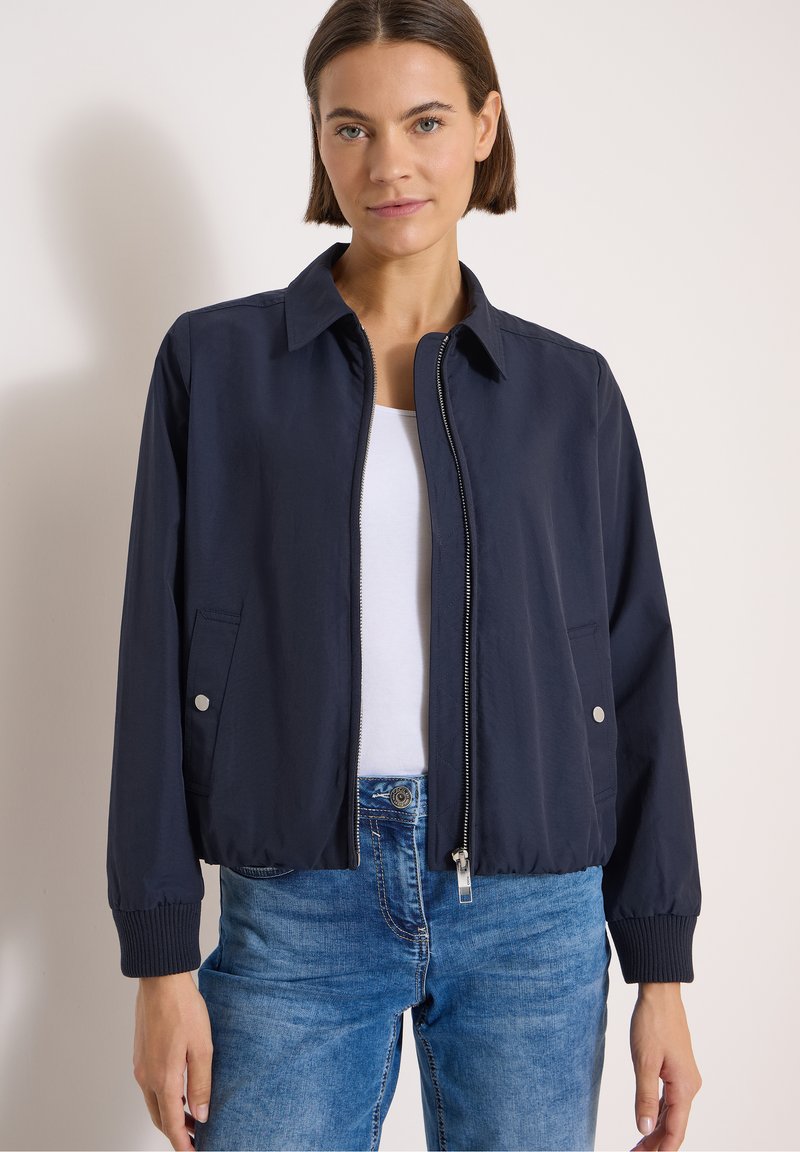 Femme portant une veste zippée bleu marine par-dessus un haut blanc et un jean bleu, debout devant un fond clair uni.