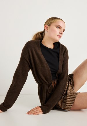 Brown knit cardigan with a tie closure, worn over a black top, paired with a patterned skirt. Smooth texture, relaxed fit, long sleeves.