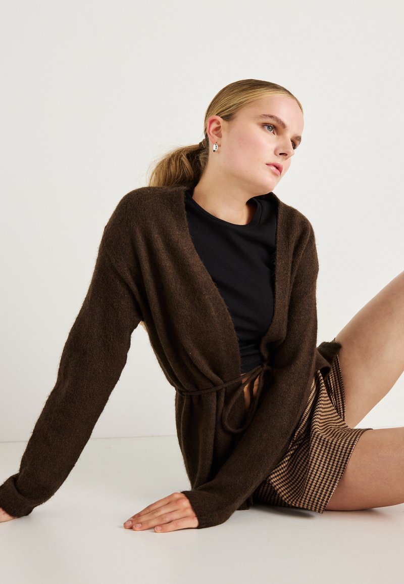 Cárdigan de punto marrón con cierre de lazo, llevado sobre una parte de arriba negra, combinado con una falda de estampado. Textura suave, corte relajado, mangas largas.