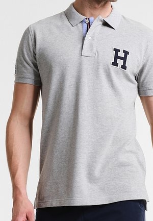 Mand iført en lysegrå kortærmet polo med en mørkeblå "H"-logo på venstre bryst og marineblå bukser.
