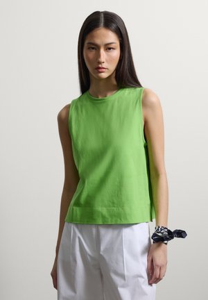 Femme portant un haut sans manches vert vif et un pantalon blanc avec un foulard noir et blanc à motif noué autour de son poignet.