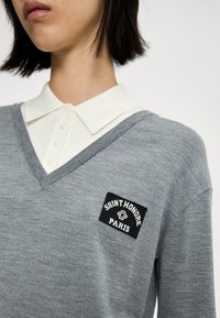 Persoon die een grijze V-hals trui draagt over een wit overhemd met kraag, met een zwarte patch op de borst waarop "Saint Honore Paris" staat.