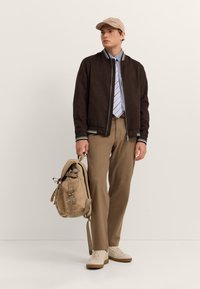 Braune Wildleder-Bomberjacke mit gestreiften Bündchen, beigefarbene Hose, gestreiftes Hemd, beiger Schirmmütze und cremefarbene Sneaker. Trägt einen braunen Rucksack.