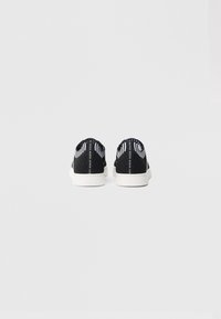 Ασπρόμαυρα sneakers χαμηλού προφίλ με λευκές σόλες, φαίνονται από πίσω, με το λογότυπο "GUESS" στις ταμπέλες στη φτέρνα και στους επάνω γιακάδες.