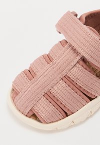 Ljusrosa tygsandal med flera vävda remmar, en texturerad mjuk sula och kardborreknäppning för justerbar passform.