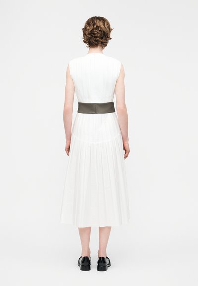 Γυναίκα με κοντά σγουρά μαλλιά, που φοράει λευκό μίντι φόρεμα χωρίς μανίκια με ευρύ σκούρο ζωνάρι και μαύρα παπούτσια, όρθια με την πλάτη στραμμένη.