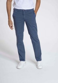 Chino bleu en coton avec une coupe slim, une taille plate et des passants de ceinture, associés à des sneakers blanches. Le tissu a une texture lisse et mate.