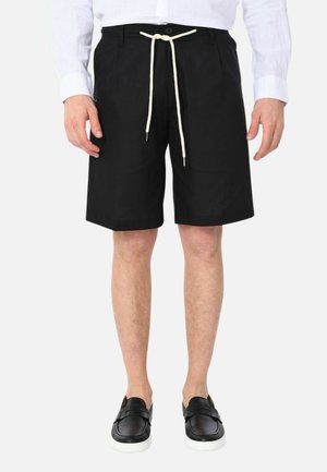 Uomo che indossa pantaloncini neri al ginocchio con coulisse bianca, camicia bianca e scarpe nere slip-on con suole bianche.