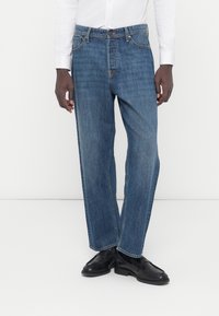 Μπλε τζιν παντελόνι με ίσια γραμμή, χαρακτηριστικά μπροστινές τσέπες, κούμπωμα με κουμπιά και διακριτικές λεπτομέρειες ραφής. Φορεμένο με μαύρα παπούτσια.