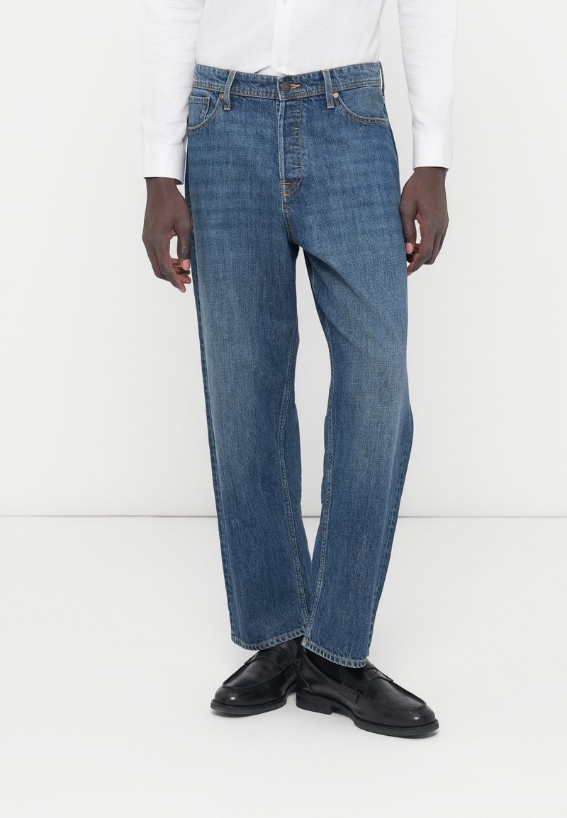 Μπλε τζιν παντελόνι με ίσια γραμμή, χαρακτηριστικά μπροστινές τσέπες, κούμπωμα με κουμπιά και διακριτικές λεπτομέρειες ραφής. Φορεμένο με μαύρα παπούτσια.