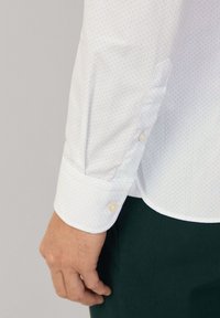 Camicia bianca a maniche lunghe con pois blu chiaro, polsini con bottoni e texture liscia. Dettagli di cucitura visibili vicino al polsino.