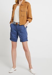 Giacca di denim marrone con tasche sul petto, camicetta bianca, pantaloncini cargo blu risvoltati e scarpe bianche slip-on, con una cintura decorata.