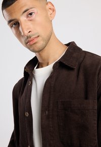 Brown corduroy shirt with a collar, front pocket, and visible buttons. Worn over a white crew neck shirt. Smooth texture and ribbed pattern.