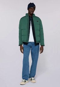 Zelená péřová bunda s vycpávkou a vysokým límcem, nošená přes bílou košili a tmavou košili na knoflíky, doplněná světle modrými džínami a teniskami.