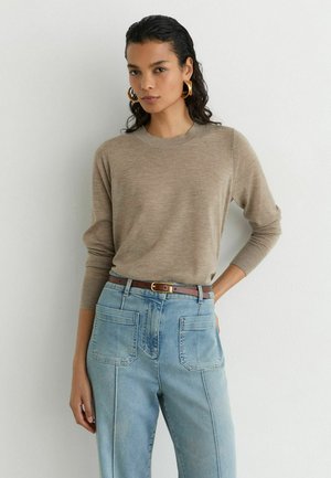 Kvinde med mørkt, vådt hår iført guldringøreringe, beige langærmet sweater, lyseblå jeans med store forlommer og brun bælte.