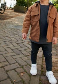 Bruine zip-up jas met twee borstzakken, grijze onderhemd, donkere broek en witte sneakers. Achtergrond van bakstenen trottoir met bladeren.