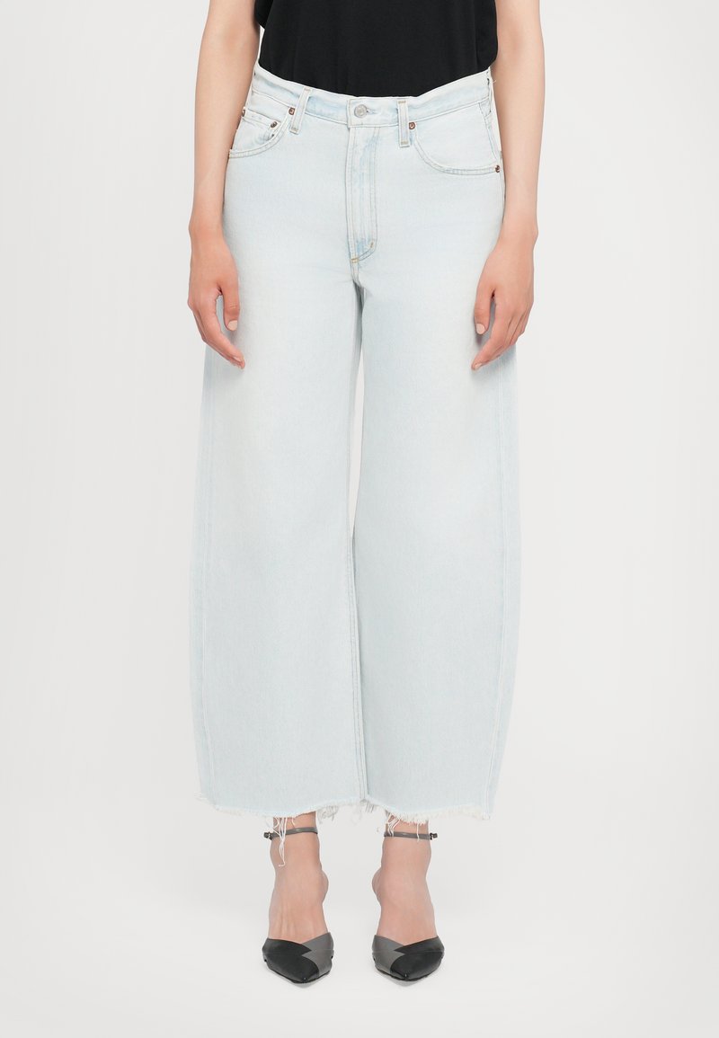 Jean large bleu clair avec taille haute, ourlets effilochés et design à cinq poches, associé à des escarpins noirs à bout pointu.