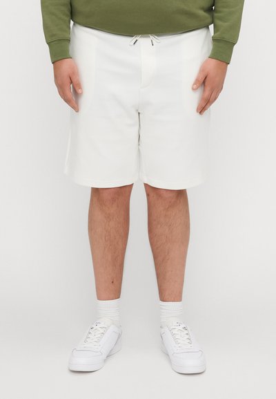 Shorts de sport blancs avec une taille élastique, dotés d'un cordon de serrage. Associés à des baskets blanches et à une chemise à manches longues verte.