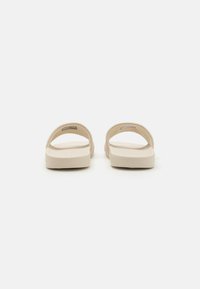 Beige natikače na slip-on s širokim trakama, udobnim uloškom i glatkom površinom. Minimalistički dizajn bez vidljivih hardverskih elemenata ili uzoraka.