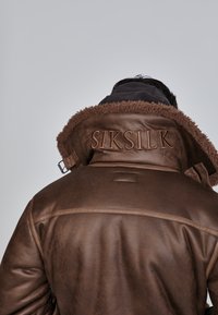 Hnedá koženka s umelou kožušinou na golieri, s vyrazeným logom "SIKSILK" na chrbte a hladkým, textúrovaným povrchom.