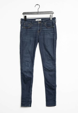Jeans skinny de mezclilla azul oscuro con una textura suave, cinco bolsillos y acentos de costura. Cuenta con un cierre de botón y pasadores para cinturón.