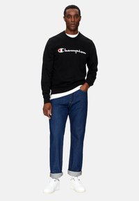 Felpa nera con logo "Champion" bianco, abbinata a jeans in denim blu e sneakers bianche. I jeans hanno i risvolti.