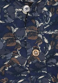 Marineblauwe bloemenoverhemd met grote crèmekleurige en bruine bloemen, gestructureerde kraag en een perzikkleurige knop met een donkere center.