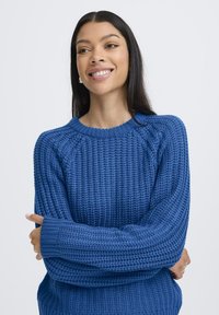 Pull en tricot bleu avec un motif texturé, col rond et manches longues. Le tissu semble épais et confortable.