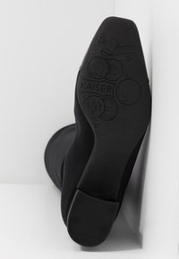 Bottes noires avec une texture lisse et élégante. La semelle présente des détails de marque embossés et des motifs similaires à des pièces de monnaie. Talon bas et plat pour plus de stabilité.