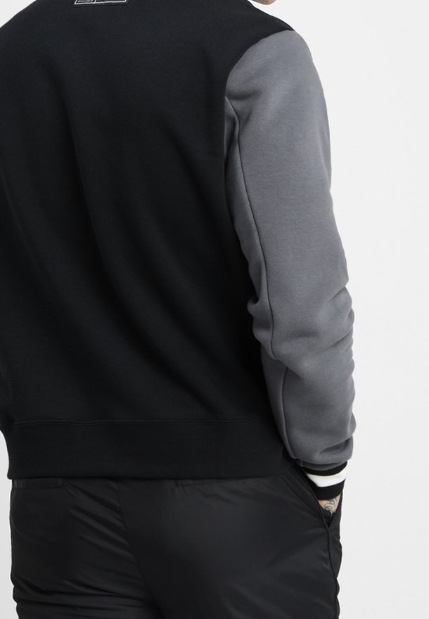 Homme portant une veste noire avec des manches grises et un pantalon noir, debout, une main dans la poche, dos à la caméra.