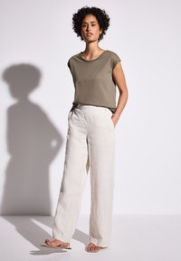 Camicia verde oliva a maniche corte abbinata a pantaloni larghi in lino color crema e sandali bassi beige, su uno sfondo chiaro.