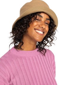 Beige bøttehat lavet af blødt stof med en bred skygge. Modellen bærer en ribbet pink sweater, viser krøllet hår og en smilende udtryk.