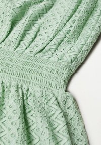 Tissu vert clair avec des motifs de dentelle texturée, doté de détails smockés à la taille et de divers motifs géométriques tout au long.