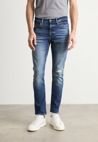 Blugi din denim albastru cu o croială obișnuită, care prezintă culori estompate, ușoare deteriorări la genunchi și un design cu cinci buzunare, asortați cu adidași albi.