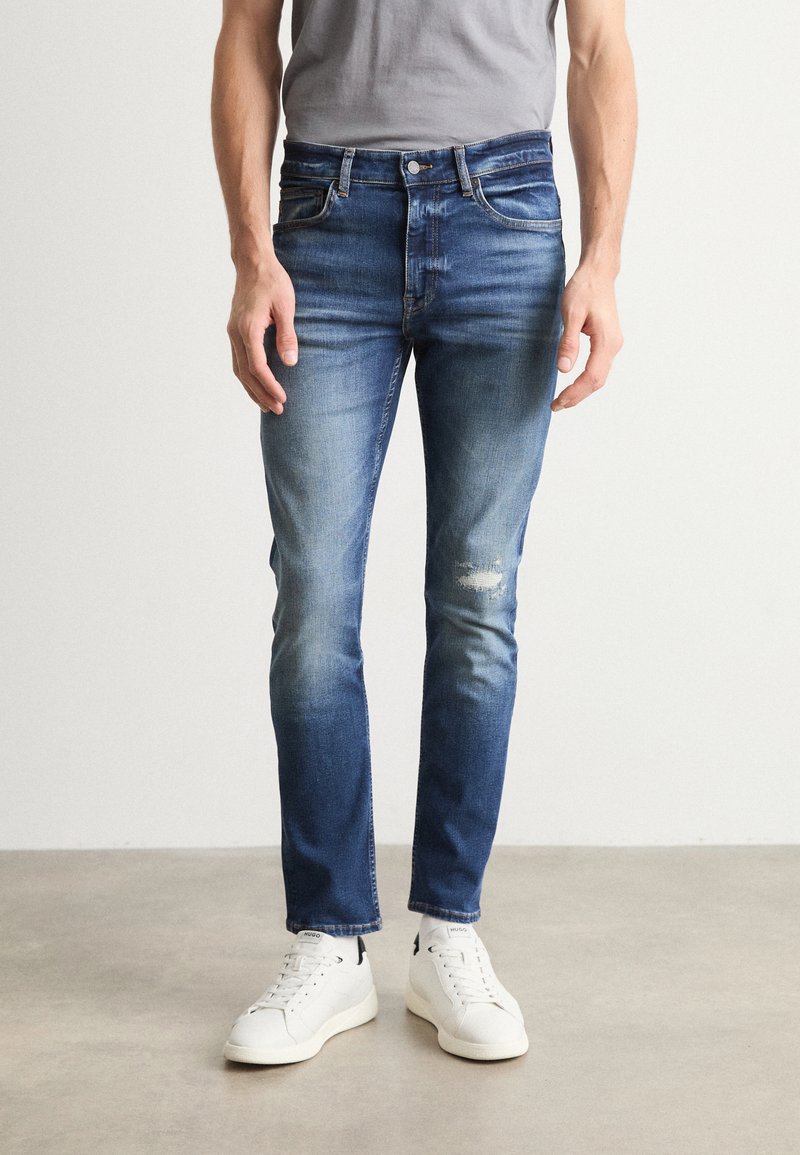 Blugi din denim albastru cu o croială obișnuită, care prezintă culori estompate, ușoare deteriorări la genunchi și un design cu cinci buzunare, asortați cu adidași albi.